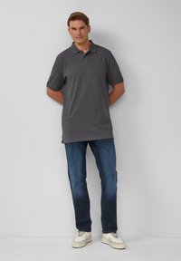 Grijze polo, korte mouwen, klassieke kraag en rechte zoom; gecombineerd met blauwe jeans en witte sneakers. Achtergrond is effen lichtgrijs.