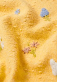 Tissu jaune avec des fleurs brodées en rose, bleu et blanc, agrémenté de petits points en relief pour la texture. Apparence douce et confortable.