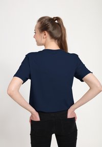 Krátký rukáv, námořnická modrá tričko s kulatým výstřihem, hladkou texturou a volným střihem, částečně zastrčené do černých džínů, pohled zezadu.