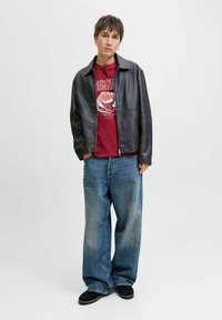 Jonge man in een zwart leren jack, rode graphic t-shirt, wijde blauwe jeans en zwarte schoenen, staand met de handen deels in de zakken.
