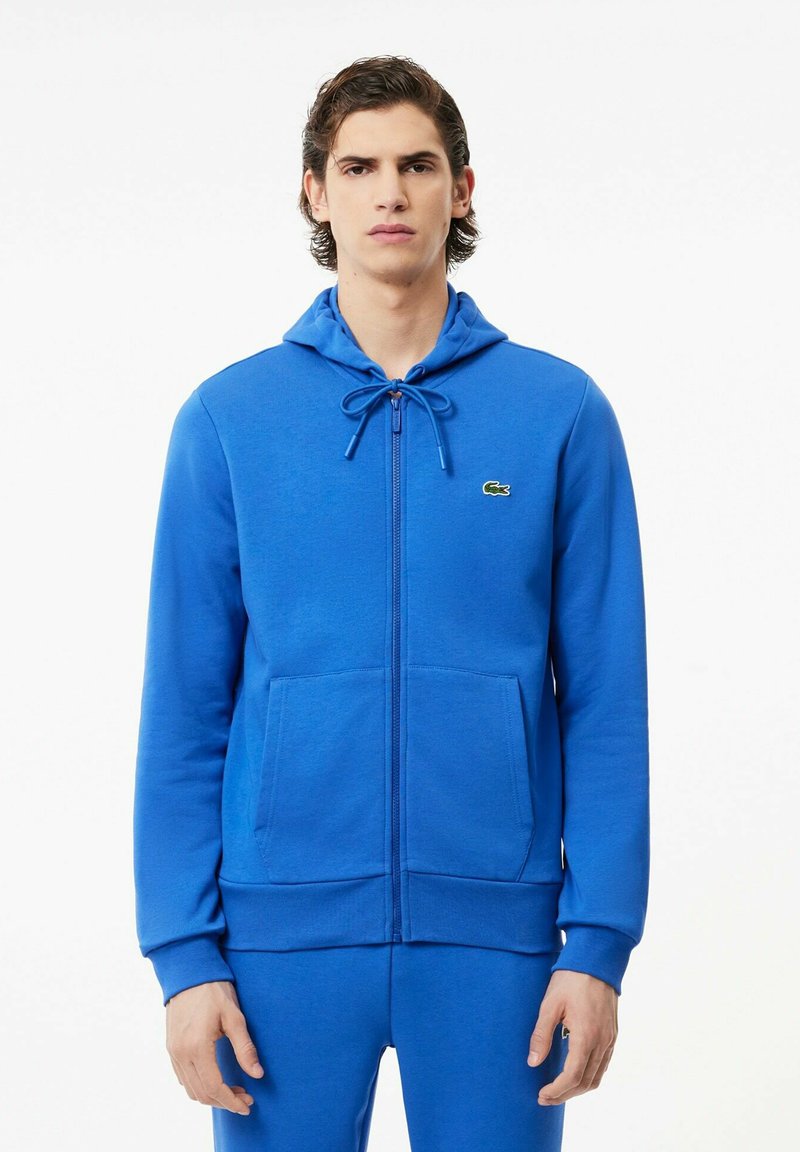 Felpa blu con zip realizzata in tessuto morbido. Presenta un cappuccio con cordino, due tasche frontali, polsini a coste e un piccolo logo sul petto.