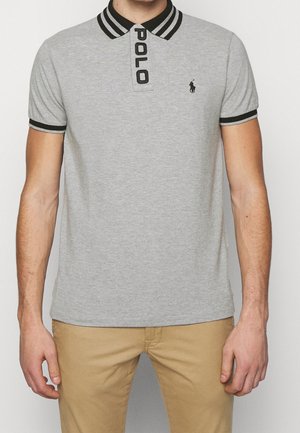 Mann trägt ein hellgraues Poloshirt mit schwarz gestreiftem Kragen und Bündchen, schwarzem gesticktem Logo und Khakihose.
