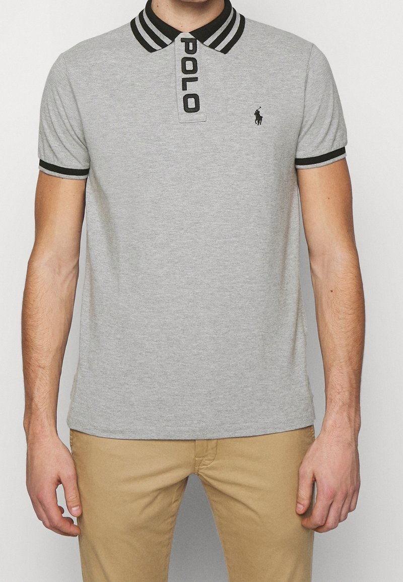 Homme portant un polo gris clair avec col et poignets rayés noirs, logo brodé noir, et pantalon kaki.