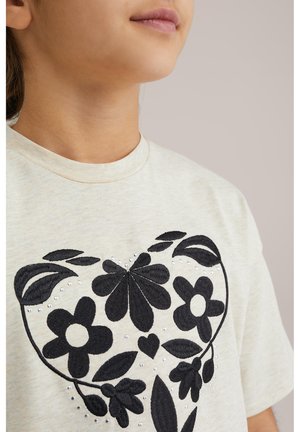 Lys beige t-shirt med korte ærmer, prydet med sorte broderede blomsterdesigns og dekorative rhinsten arrangeret i en hjerteform.