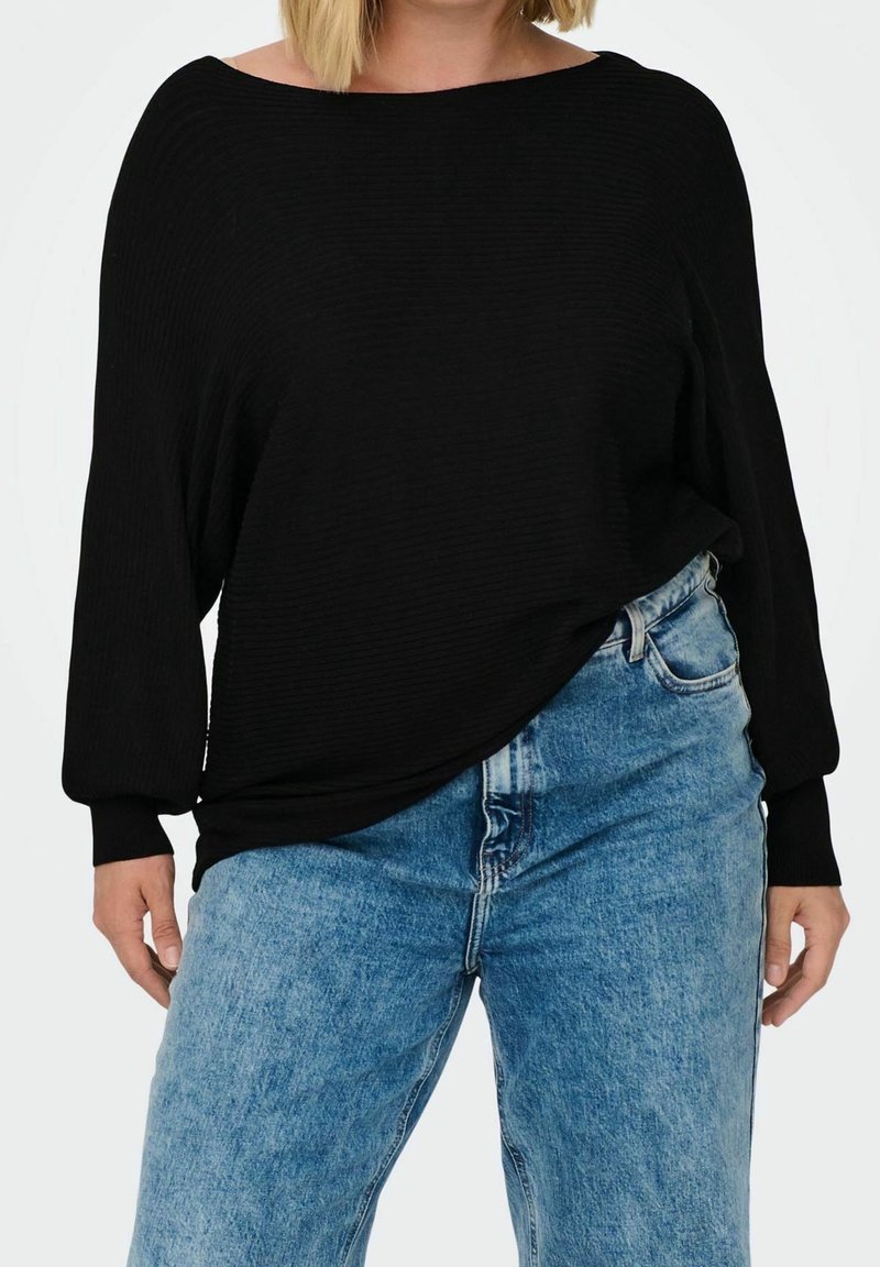 En kvinna med blont hår bär en lös svart topp och ljusblå högmidjade jeans, stående mot en enkel bakgrund.