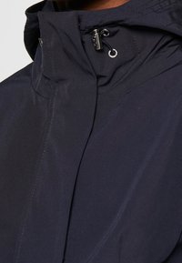 Marinblå jacka med hög krage, stilren design och dragsko-stängning. Tillverkad av ett slätt, lättviktigt material med små sömdetaljer.