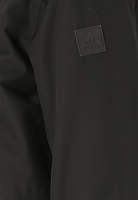 Veste imperméable noire au toucher lisse, avec un patch en caoutchouc sur la manche et des éléments de design minimalistes.