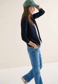 Jeune femme en veste bleu marine, jean bleu, casquette verte et baskets blanches, marchant avec une main dans la poche et souriant.