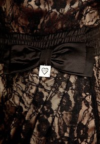 Abito in pizzo nero con motivi floreali, caratterizzato da una fascia in vita plissettata e un fiocco nero in satin, impreziosito da un ciondolo argentato a forma di cuore.