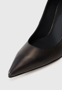 Pantof de piele neagră cu toc înalt, având vârful ascuțit și părți laterale decupate, prezentând o textură netedă și un design elegant.