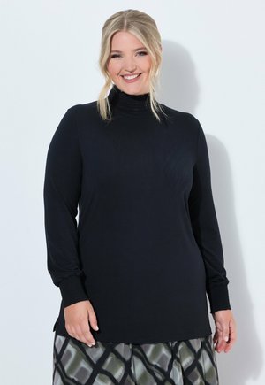Lächelnde Frau mit blondem Haar trägt ein schwarzes Langarmshirt mit Manschetten und einen gemusterten Rock, sie steht vor einem schlichten hellen Hintergrund.