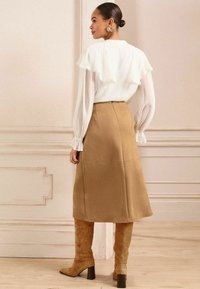 Blusa bianca con dettagli a volant e maniche trasparenti abbinata a una gonna a trapezio khaki e stivali in suede color cammello al ginocchio con tacco a blocco.