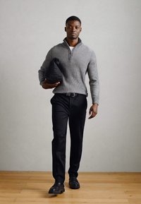 Suéter gris de canalé con media cremallera, pantalones negros y zapatos negros. El modelo sostiene un bolso negro, de pie sobre un suelo de madera frente a un fondo neutro.