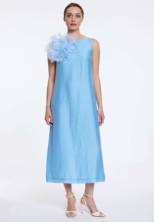 Femme portant une robe midi sans manches bleu clair avec un grand détail de fleur volantée sur une épaule et des sandales blanches à talons hauts.