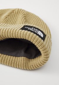 Strikket lue i beige med en svart logo-patch. Har ribbestruktur og en brettet oppkant, som viser en mørk innside.