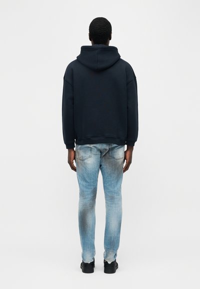 Άνδρας που φοράει μαύρο φούτερ με κουκούλα και ανοιχτό γαλάζιο ξεθωριασμένο τζιν, στέκεται γυρισμένος με την πλάτη σε απλό λευκό φόντο.