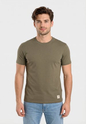 Joven con cabello corto y ondulado de color castaño, vistiendo una camiseta de cuello redondo verde oliva y jeans azul claro, de pie contra un fondo liso.