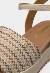 Κοντινό πλάνο μπεζ και καφέ υφασμένης σανδάλιας με λουράκι στον αστράγαλο και χρυσό λογότυπο "W" στο μπροστινό λουράκι.