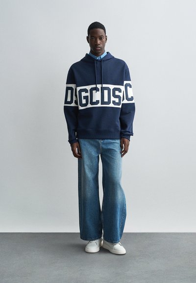 Fiatal férfi áll, sötétkék kapucnis sweatshirtöt visel fehér logós csíkkal, laza, széles szárú kék farmernadrágot, és fehér sportcipőt, egyszerű világosszürke háttér előtt.