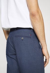 Pantalones a cuadros azules con un bolsillo trasero, cierre de botón y pasadores para cinturón, que presentan un pequeño detalle de logo en rojo, blanco y azul.