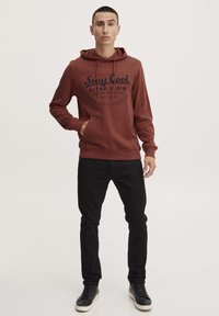 Felpa color ruggine con tasca frontale, caratterizzata da testo nero e cordini. Abbinata a jeans neri e sneakers nere.