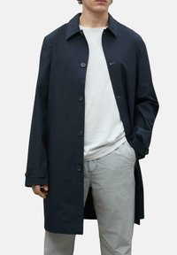 Trinchera navy hecha de tela suave, con un collar clásico, cierre de botones y bolsillos laterales, combinada con una camiseta ligera y pantalones.