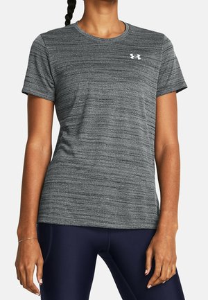 Kobieta ubrana w dopasowany szary sportowy t-shirt z krótkim rękawem z białym logo Under Armour oraz ciemnogranatowe legginsy, stojąca z rękami swobodnie opuszczonymi.