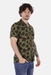 Camicia a maniche corte di colore verde oliva con un motivo a palme nere, chiusura con bottoni e colletto rilassato. Tessuto leggero.