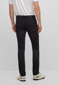 Uomo in jeans neri slim-fit, t-shirt bianca e sneakers bianche, in piedi contro uno sfondo grigio chiaro uniforme, vista da dietro.