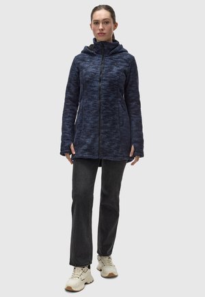 Giacca in pile blu navy con zip frontale, colletto alto e cappuccio. Tessuto testurizzato con due tasche laterali. Indossata con jeans scuri e sneakers chiare.