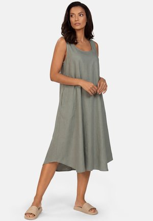 Kobieta w szarej, luźnej, bezrękawnikowej sukience midi i beżowych sandałach typu slide, stojąca z jedną nogą lekko wysuniętą do przodu na jednolitym tle.