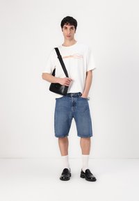 White graphic t-shirt with orange text, denim shorts, black crossbody bag, white socks, and black loafers. Simple design, casual fit.