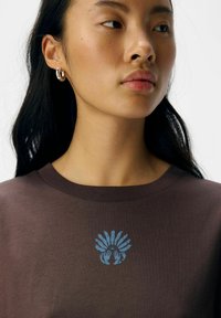 Bruine T-shirt met korte mouwen, versierd met een blauw grafisch ontwerp van handen op de borst. Zachte stof met een klassieke pasvorm en ronde hals.