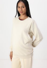 Sudadera beige con un escote redondo y puños acanalados. Presenta un diseño de corte lateral, confeccionada en una tela suave, llevada sobre una blusa de color burdeos.