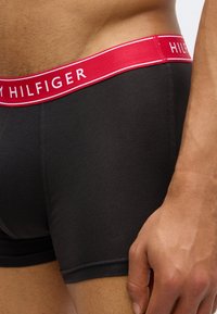 Close-up of a person wearing black boxer briefs with a red waistband displaying the Tommy Hilfiger brand name.