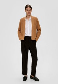 Hellbrauner gerippter Cardigan über einem weißen Hemd, kombiniert mit schwarzen Hosen und schwarzen Slippern. Das Ensemble zeichnet sich durch einen lässigen Schnitt und klare Linien aus.