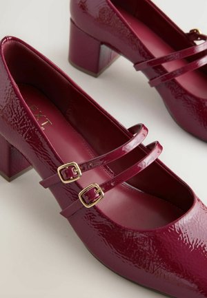 Burgundy patent leather heels with a square toe, two straps featuring gold buckles, and a block heel. Smooth texture and glossy finish.