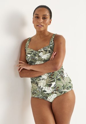 Vrouw met gekruiste armen, gekleed in een badpak met een patroon van groene, witte en bruine tropische bladeren, staande tegen een effen witte muur.