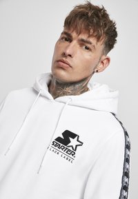 Hanorac alb cu șnur, având un logo brodat în negru "Starter Black Label" pe piept și dungi laterale negre cu un model.