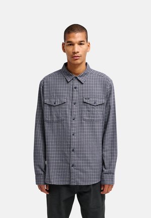 Homme portant une chemise à carreaux grise et bleue à boutons avec deux poches poitrine, debout devant un fond blanc uni.