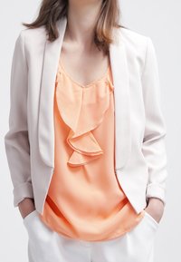 Blazer i lys beige stof over en ferskenfarvet bluse med flæser. Blusen har en blød texture; outfittet er parret med hvide bukser.