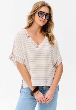 Kobieta o falowanych blond włosach nosi duże okulary przeciwsłoneczne w oprawkach w kolorze żółwia, luźną beżową bluzkę w paski z dekoltem w kształcie litery V, jasnoniebieskie dżinsy i bransoletki.