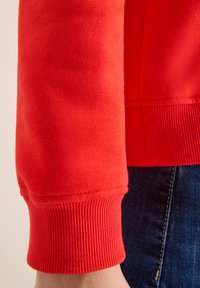 Sudadera roja con puños acanalados y un tejido suave y texturizado. Vista en primer plano que muestra el detalle de la manga y la costura.