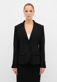 Blazer nero su misura con rever a notched, chiusura a un bottone, due tasche frontali e dettagli con bottone sulle maniche. Texture liscia.