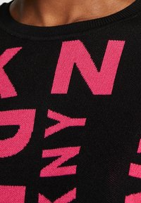 Pull en maille noire avec un grand texte rose en caractères gras. Encolure arrondie avec bord côte. Vue en gros plan montrant la texture et le contraste des couleurs.