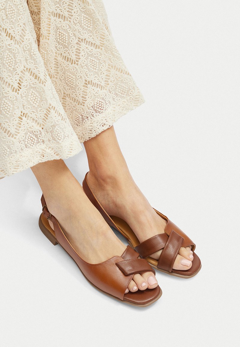 Sandalias de cuña de cuero marrón con la punta abierta, que presentan un diseño de bandas cruzadas y una suela plana. La textura parece lisa con un detalle minimalista.