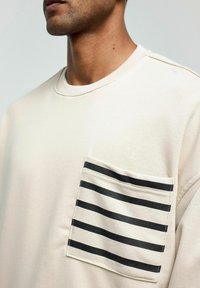 Beige sweatshirt met een ronde hals, voorzien van een linkerschouderzak met horizontale zwart-witte strepen en een zachte, textuurlichte afwerking.