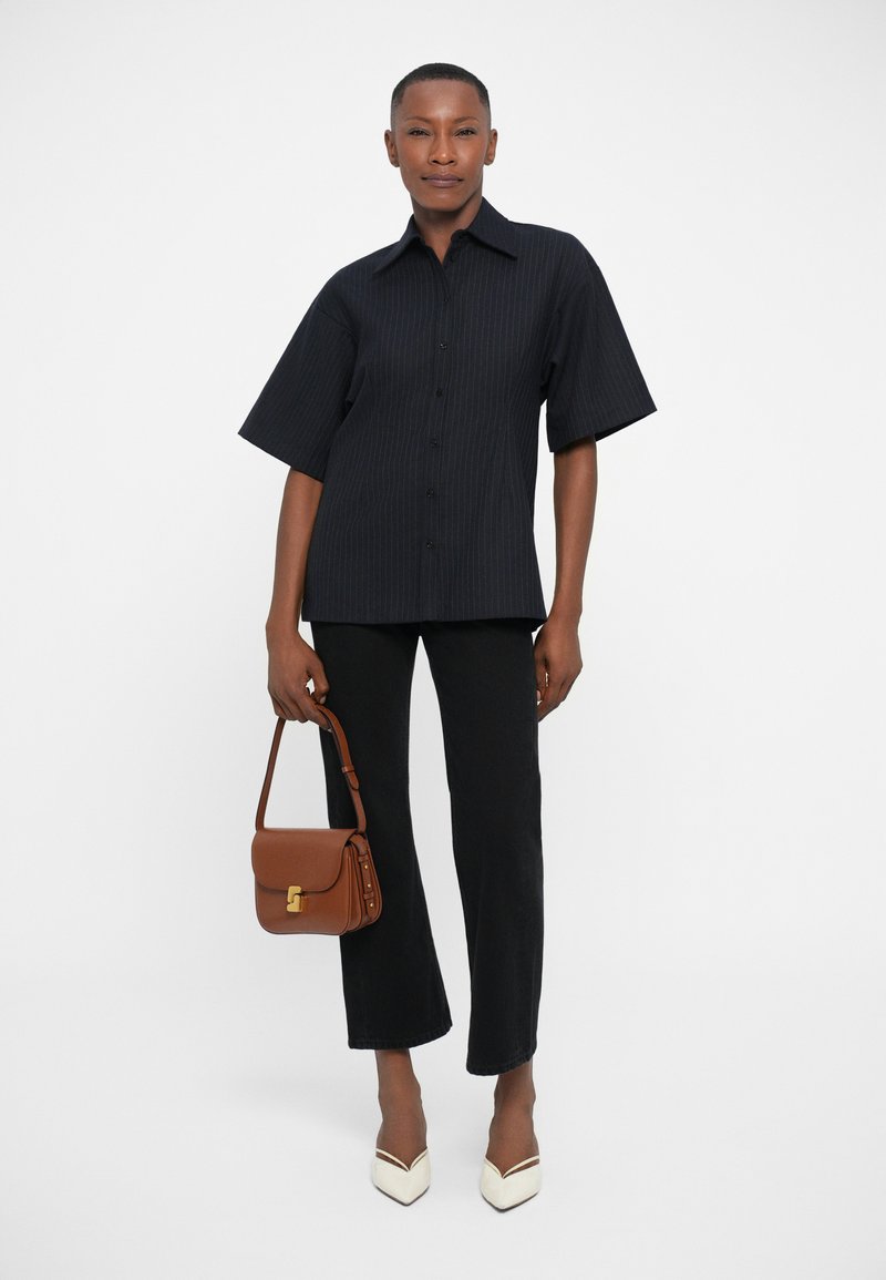 Chemise noire à rayures, pantalon noir évasé, escarpins blancs à bout pointu, et sac en bandoulière marron avec un fermoir doré.