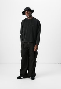 Zwart long sleeve shirt met een ronde hals; losse zwarte cargobroek met meerdere zakken; zwarte emmerhoed; zwarte schoenen.