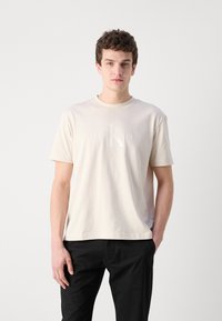 Light beige cotton t-shirt with round neckline and short sleeves. Features an embossed Calvin Klein logo on the chest. Paired with black pants.